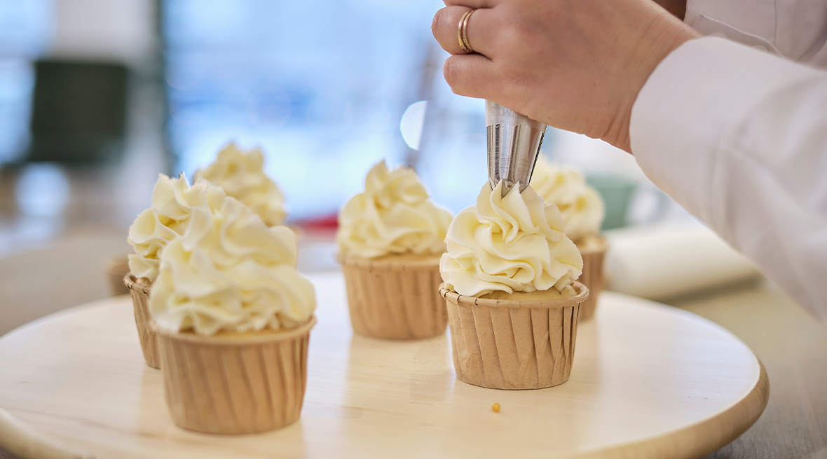 Whipped cream being squeezed out of a piping bag onto a cup cake, with several other cakes on a round board